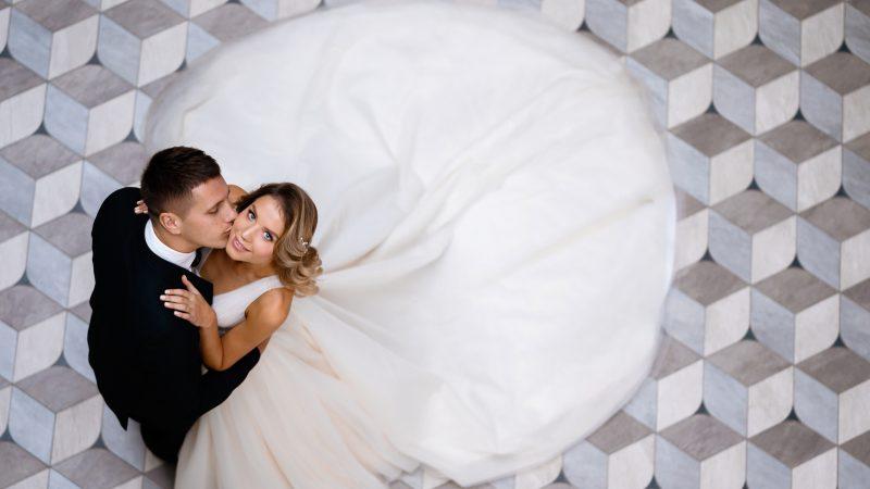 Above view of stylish and serious bridegroom, wearing in black suit, standing and kissing his wife. Attractive and cheerful bride in white gown with long train, bending body on aback and embracing her husband, looking up while posing on geometric floor tile indoor